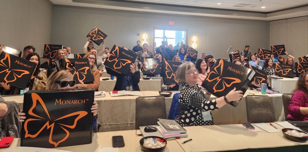 A room full of people sitting in rows at long rectangular tables smile as they hold up their black Monarch boxes, which prominently feature a large outline of an orange butterfly outline and the Monarch logo.