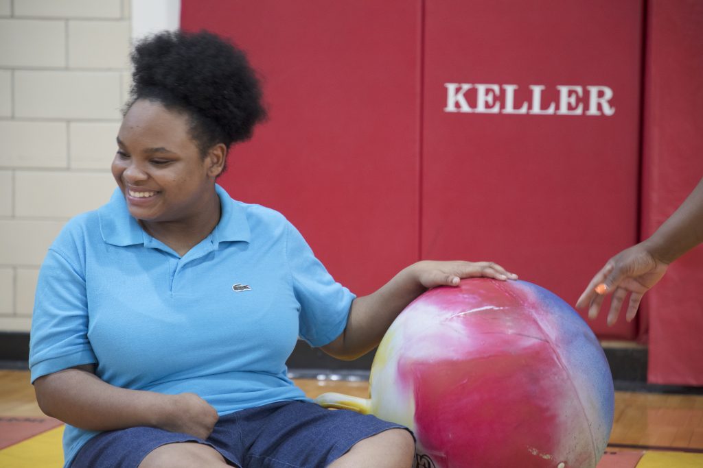 A smiling older girl wearing a bright blue polo shirt holds a colorful ball as someone else’s hand reaches for it. Behind her, a red wall with the word “KELLER” printed on it can be seen.