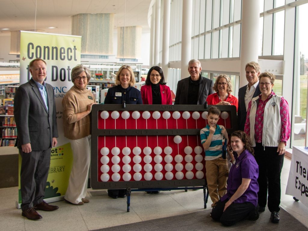 A group of eight adults pose for a photo while standing around a gigantic cranmer abacus. Another adult squats with a young child near the bottom right corner of the abacus. Behind the two leftmost standing adults, a tall thin banner reading “Connect the Dots” can be seen.