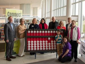 A group of eight adults pose for a photo while standing around a gigantic cranmer abacus. Another adult squats with a young child near the bottom right corner of the abacus. Behind the two leftmost standing adults, a tall thin banner reading “Connect the Dots” can be seen.