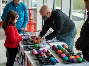 An adult speaks to a young girl from across a table while another man stands at the head of the table. The table between the adults and the child has several muffin tins filled with colorful tennis balls.