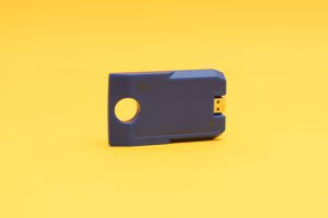 Angled front view of the blue digital talking book cartridge against a yellow background. The cartridge has a circular hole on one end and a USB port on the other, and it reads 