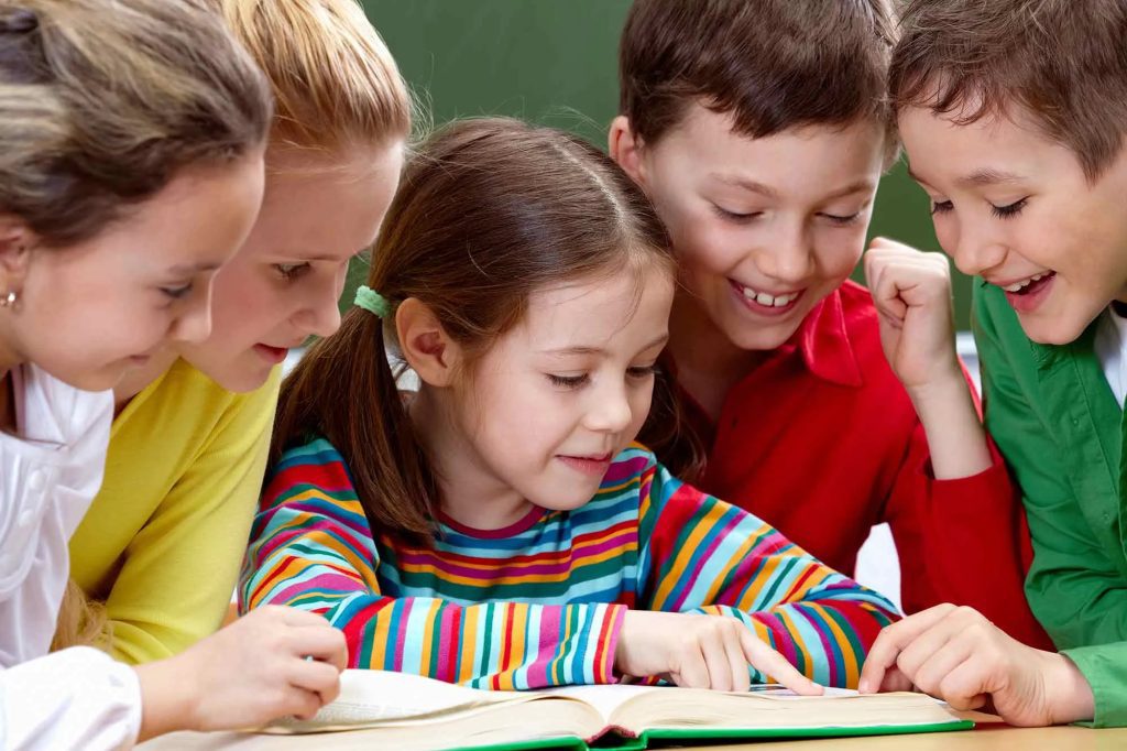 Five children with excited expressions are gathered around a book. The child in the center and the child on the far right are each touching the book with one of their index fingers.