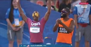 A blindfolded man wearing a USA athletic shirt and a runner’s bib with the name “BROWN” on it raises his arms high in a victorious stance as the man standing beside him, who is wearing a bright orange shirt with the word “GUIDE” on it, claps. The two men appear to be standing in a stadium. A small pop up at the bottom of the image reads “3, USA, David R Brown - T11” with a small American flag by the word “USA.”