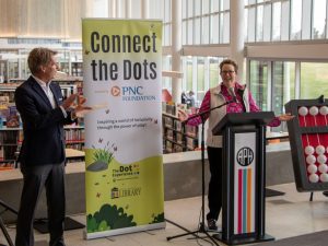 A woman animatedly speaks from behind a podium with the APH logo on it. To her right, a man is clapping, and between her and the man is a tall thin banner reading “Connect the Dots powered by PNC Foundation. Inspiring a world of inclusivity through the power of play!” over The Dot Experience logo and the Louisville Public Library logo.