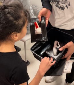 An adult hands a child a STACS card with the tip of a white cane on it. The adult is also holding a small work-play tray containing another STACS card with a small shoe and a label reading "WALK" on it.