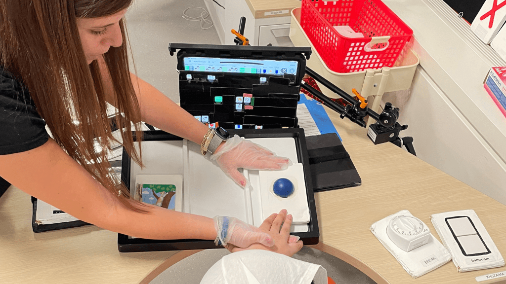 A woman wearing plastic gloves guides a child's hand to a STACS card with a blue ball on it. Other STACS cards can be seen on the table they are using.