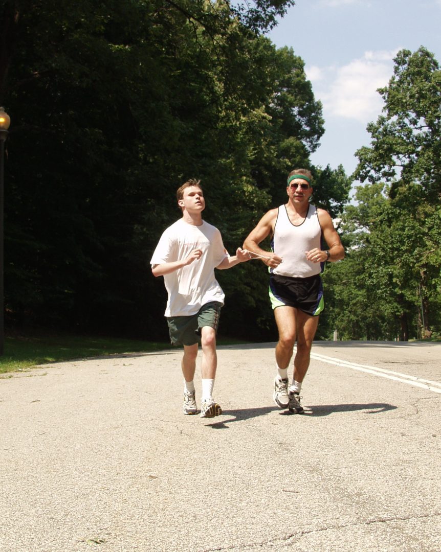 An adult man and a teenage boy wearing running gear and attached at the hands by a short teather jog on a road surrounded by trees on a sunny day.