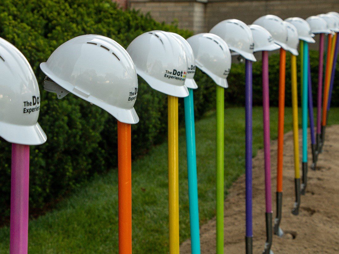A line of shovels with colorful handles stuck in the dirt. On top of each handle is a white construction helmet with The Dot Experience logo.