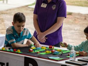 A boy plays with tactile town, which is set up on a table while an adult in a purple shirt stands beside him.