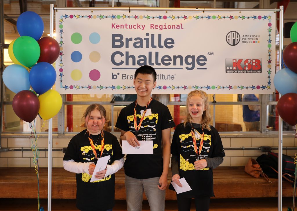 Three students wearing medals and holding white envelopes smile as they stand in front of a Kentucky Regional Braille Challenge banner, which is flanked by colorful balloons on both sides.