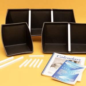 All components of the Black Expandable Calendar Boxes kit laid out on a yellow background. Included are six black foam boxes attached to one another with white U-channels, two additional large white U-channels, six smaller u-Channels, and the print guidebooks placed in front.