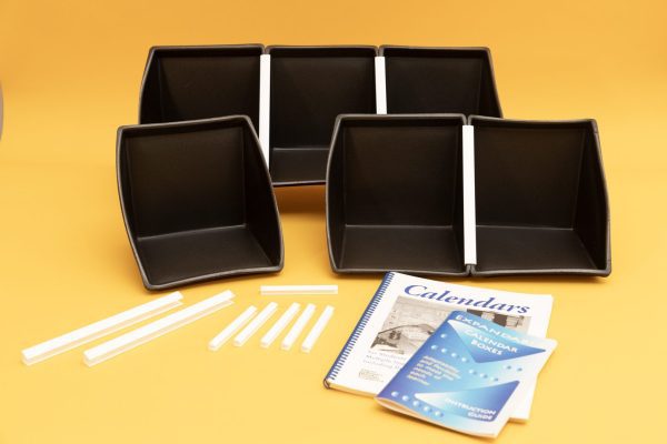 All components of the Black Expandable Calendar Boxes kit laid out on a yellow background. Included are six black foam boxes attached to one another with white U-channels, two additional large white U-channels, six smaller u-Channels, and the print guidebooks placed in front.