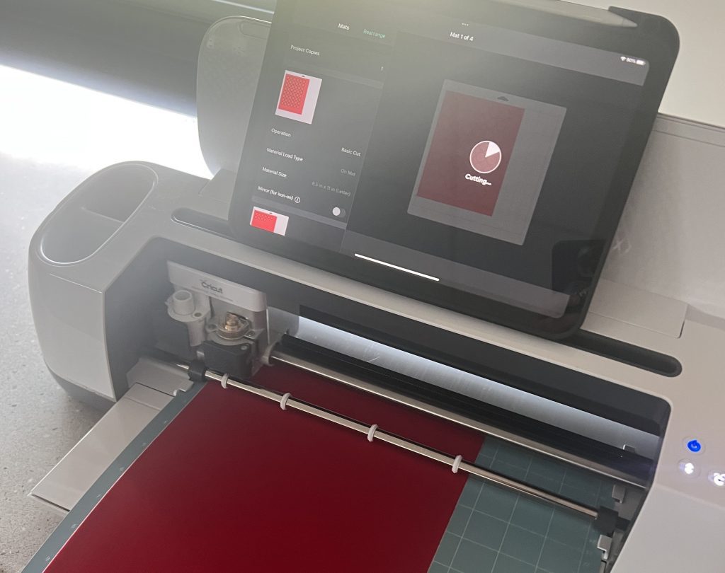 A silver Cricut machine, which looks like a printer with a tablet attached to the top and a cutting board with measurements where the printer tray would be, cutting a large red rectangular piece of material.