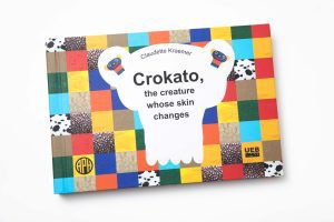 The cover of LDQR's Crokato, the Creature Whose Skin Changes by Claudette Kraemer. The cover has a checkerboard pattern and features the APH and UEB logos.