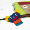 : The removable felt character Crokato lays between two open pages, one containing a tactile illustrated flowerpot. Crokato is blue with yellow ears, a silver nose, and an orange shirt.