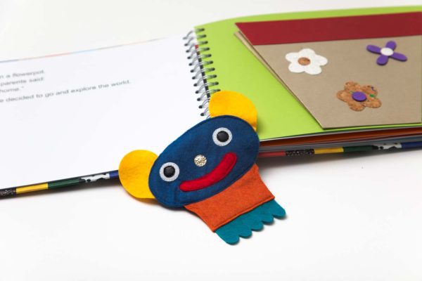 : The removable felt character Crokato lays between two open pages, one containing a tactile illustrated flowerpot. Crokato is blue with yellow ears, a silver nose, and an orange shirt.