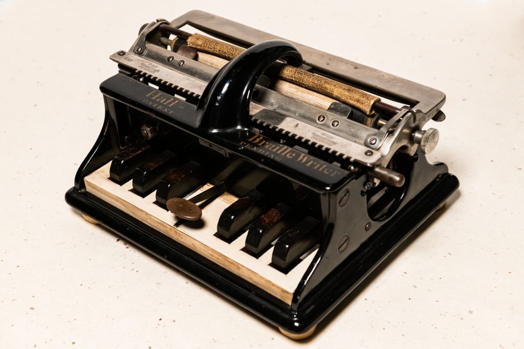 An original Hall Braille Writer with a glossy black and silver metal exterior and glossy black keys with a white surface under them. The words “Hall Braille Writer Patent Pending” sit above the keys and are separated by the braille writer’s handle. A worn cylindrical piece of leather can be seen wrapped around one of the horizontal metal rods toward the back of the braille writer.