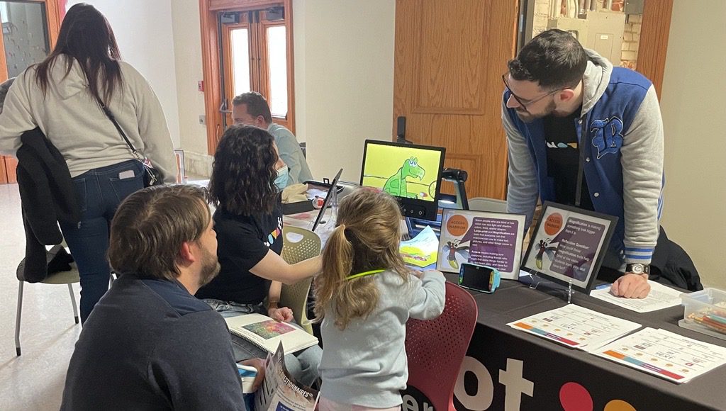 A standing man and a seated woman show a seated young girl and a kneeling man a colorful image of a cartoon dinosaur on the Jupiter’s screen.