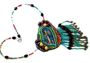 An ornate beaded talisman lies on a white surface. It is adorned with onyx, seafoam green and pink stones, and many other cool-hued beads and stones. The beads cascade down in thin tassels from the bottom of the talisman, and a beaded wire connects to both of its upper corners.