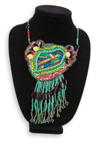 An ornate beaded talisman rests gently on a black neck mannequin. It is adorned with onyx, seafoam green and pink stones, and many other cool-hued beads and stones. The beads cascade down in thin tassels from the bottom of the talisman, and a beaded wire connects to both of its upper corners.