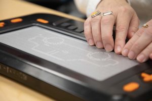 Two hands wearing several rings feel a tactile graphic of a car displayed on the Monarch’s 10-line by 32-cell refreshable braille display.