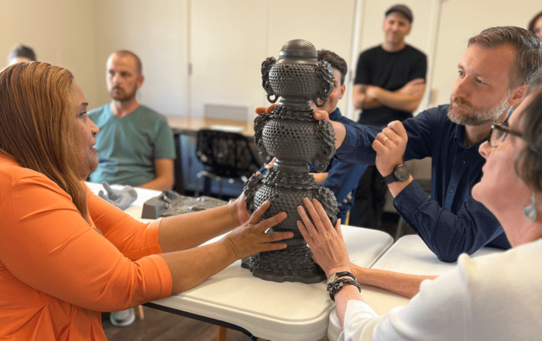 Four seated people touch a charcoal-colored protoype of an intricate incense burner, which is standing on a table. Several people in the background look on.