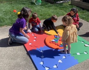 Two APH staff members teach a group of three small children about shapes and colors with the APH Reach and Match Kit. The mat is red, blue, yellow, and green. The children are on top of the mat exploring the various shapes.