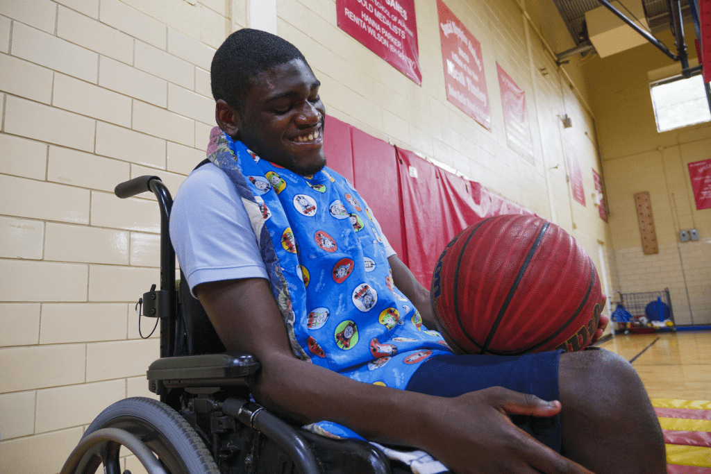 A teenage boy sitting in a wheelchair smiles as he holds a basketball in a gymnasium.