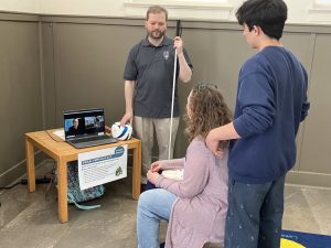 A man holding a white cane stands beside a computer, which is displaying a person’s face while sitting on a low table. In front of the computer, a woman is sitting in a chiar while a boy stands behind her.