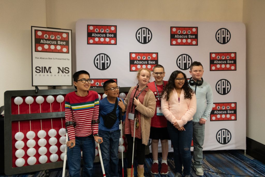 Six students of various races and ages standing in front of a giant abacus and a backdrop with the APH, Abacus Bee, and Simons Foundation logos.