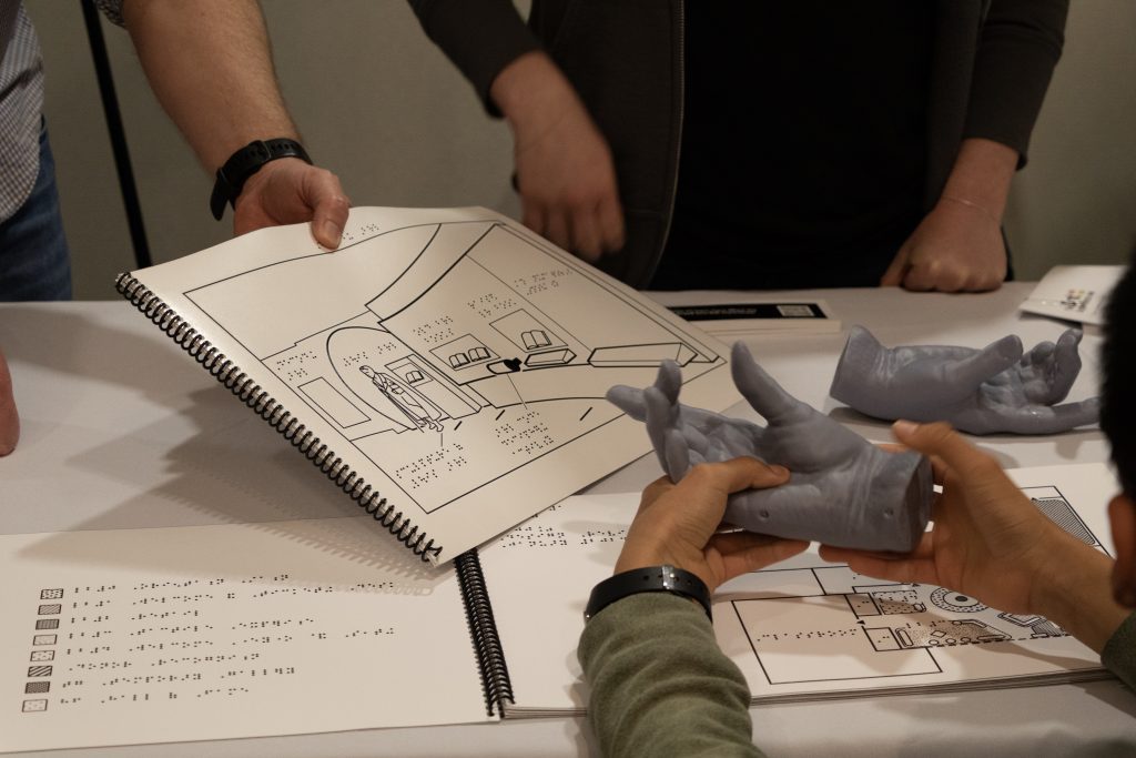 A child holds a grey mold of a hand in their hands over a table while a person on the other side of the table holds out a black and white line drawing of a museum exhibit with braille labels. On the table, another mold of a hand and a thin spiral bound book open to pages showing a key in braille and a floor layout diagram are visible. A second person can be seen behind the table.