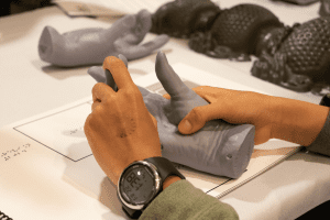 Two human hands holding a grey prototype hand. Another hand prototype and a prototype of an intricate inscence burner can be seen in the background.