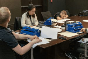 Four students, who all appear very focused, sit around a long wooden table covered in braille paper, lined paper, Perkins Braillers, and abacuses. 