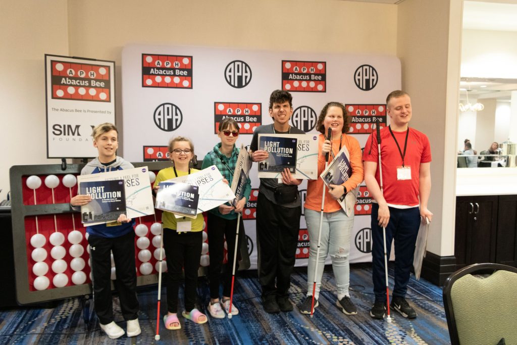 Six students of various races and ages standing in front of a giant abacus and a backdrop with the APH, Abacus Bee, and Simons Foundation logos. Some students are holding tactile books about STEM and space.