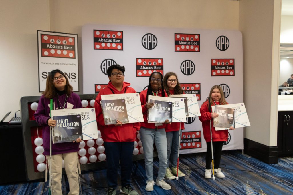 Five students of various races and ages standing in front of a giant abacus and a backdrop with the APH, Abacus Bee, and Simons Foundation logos. Some students are holding tactile books about STEM and space.