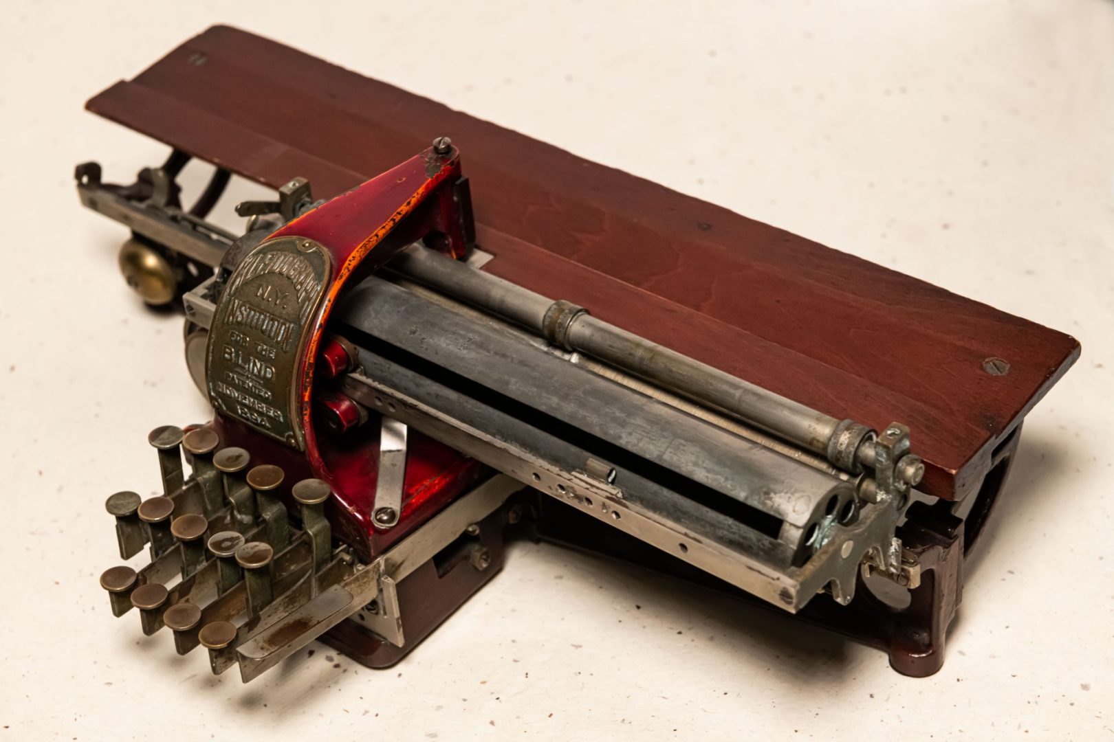 A Kleidograph with metal keys, a reddish wood back, and a metal plate indicating that the braille writer belongs to the "N.Y. Institution for the Blind."