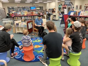 A group of children and their parents sit on colorful stools in a library's reading area as a man who is blind reads a braille storybook.