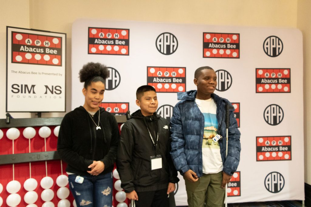 Three students of various races and ages standing in front of a giant abacus and a backdrop with the APH, Abacus Bee, and Simons Foundation logos.