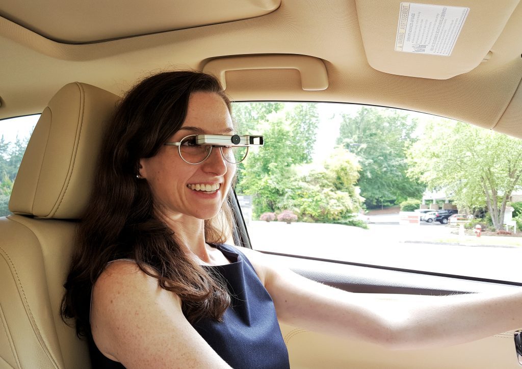 A woman wearing bioptic lenses smiles as she sits in the driver's seat of a car.