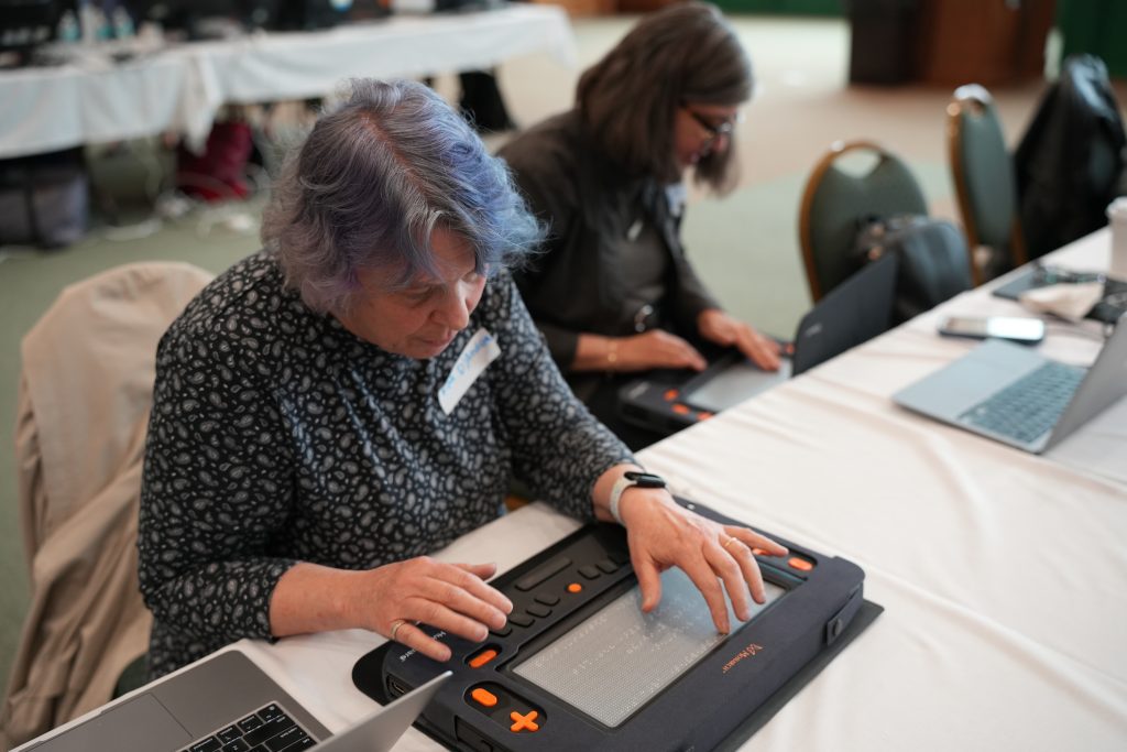 A woman seated at a table leans over a Monarch as she touches braille on its multiline braille display. Another woman using the Monarch on her lap with a laptop on the table in front of her can be seen in the background.