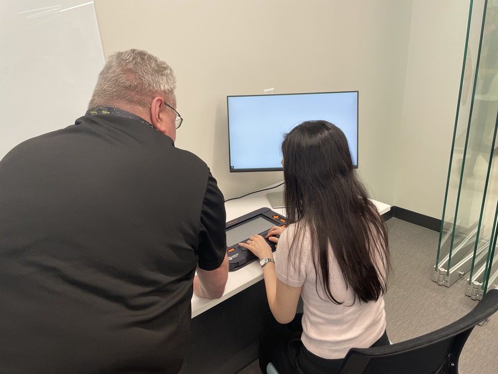 A man leans down to help a seated young woman use the Monarch, which is laying on a table in front of her. A computer monitor can be seen to the right of the Monarch.