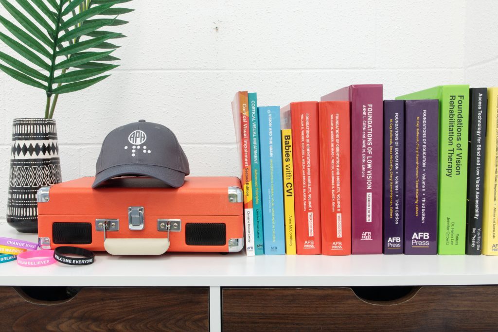 On the top of a bookshelf, a grey APH baseball cap sits on an small orange briefcase with a line of APH Press titles to the right and a pile of rubber braille APH bracelets and a decorative plant in a small vase to the left.