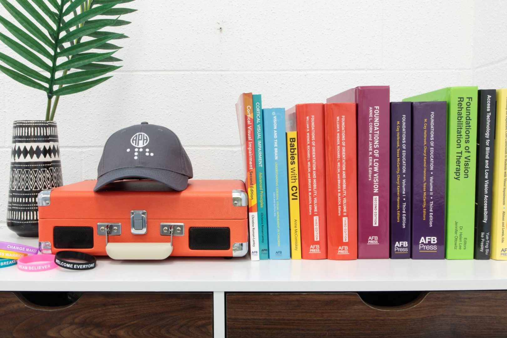 On the top of a bookshelf, a grey APH baseball cap sits on an small orange briefcase with a line of APH Press titles to the right and a pile of rubber braille APH bracelets and a decorative plant in a small vase to the left.