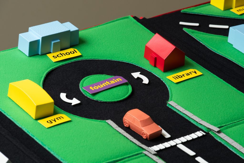 A close up view of Tactile Town with a labeled library, a school, and a gym visible on the outside of a roundabout that an orange car is exiting.