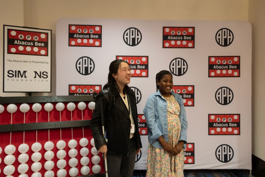 Two students of various races and ages standing in front of a giant abacus and a backdrop with the APH, Abacus Bee, and Simons Foundation logos.