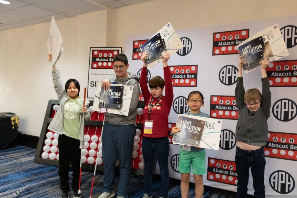 Five students of various races and ages standing in front of a giant abacus and a backdrop with the APH, Abacus Bee, and Simons Foundation logos. Some students are holding tactile books about STEM and space. Three of the students have their books raised above their heads.