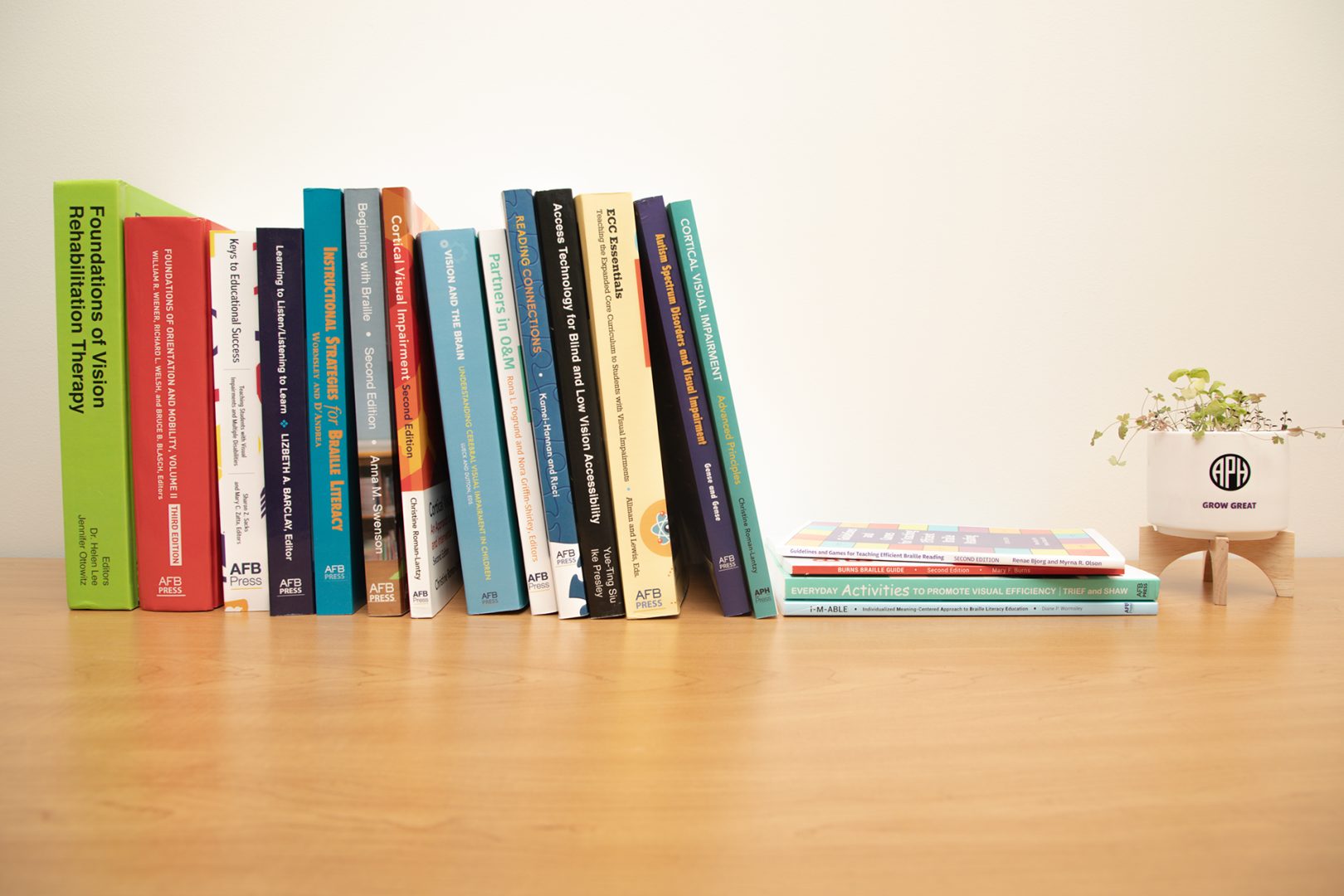 On a wooden surface against a white wall, several APH Press titles are stacked horizontally, including Foundations of Vision Rehabilitation Therapy, Cortical Visual Impairment, and ECC Essentials among many more! To the right of these is a short stack of vertically arranged titles. Next to those is a small white planter with an APH logo on a wooden stand containing a green sprouting plant.