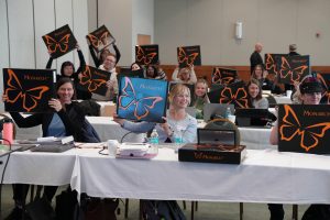 Four rows of smiling people, most sitting behind long tables each covered by a white tablecloth, and a few standing, hold their Monarch boxes up.