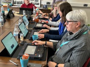 Five people seated on one side of a wooden conference table, each with a Monarch refreshable braille displays and a small monitor in front of them. An additional person who also has a Monarch and a monitor is seated at the head of the table.